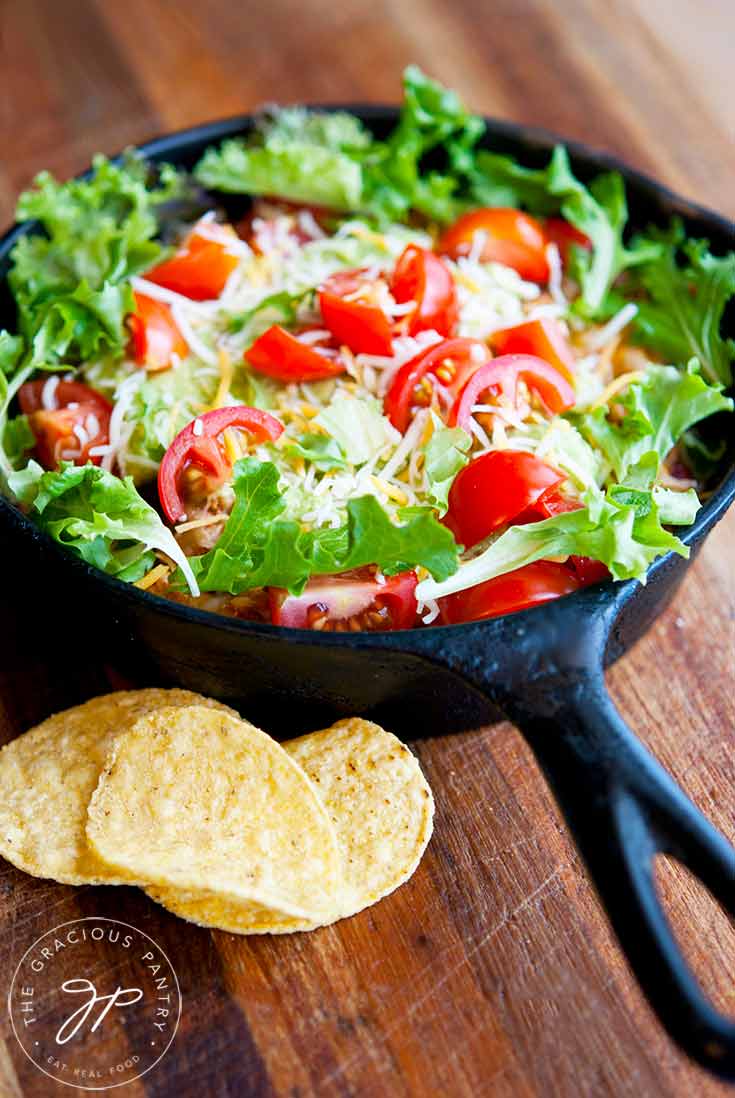 A small, cast iron skillet sits on a wood surface, filled with this Clean Eating Skillet Taco Salad. You can see beautiful, bright green lettuce and fresh-cut, red cherry tomatoes with shredded cheese sprinkled over the top. A few corn chips sit to the side.