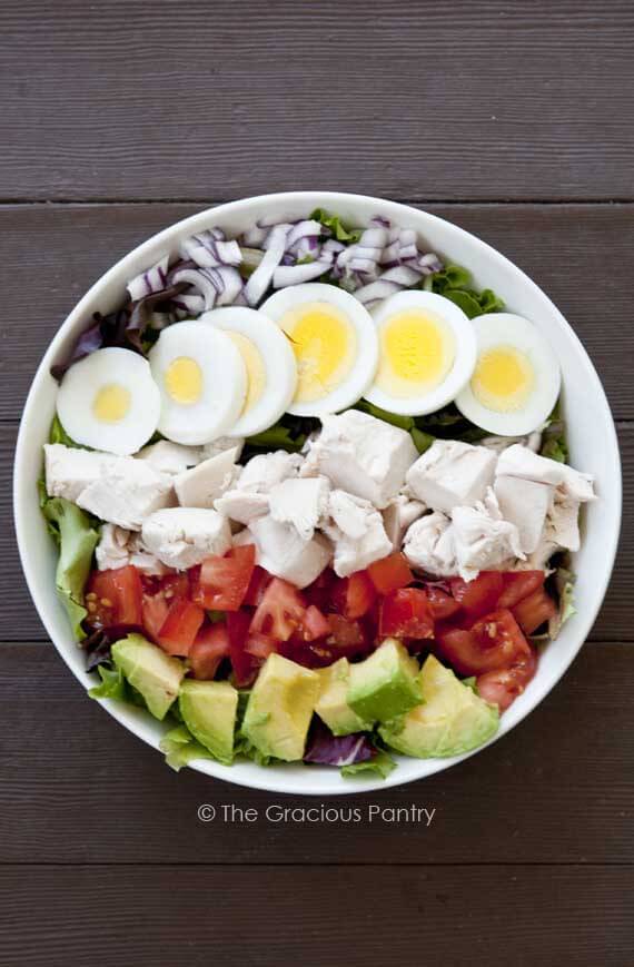 A Clean Eating Cobb Salad in a white bowl on a dark wood background. The salad is shown from over head, looking down into the bowl. The salad is unmixed so you can see each of the ingredients arranged nicely on top of the lettuce.