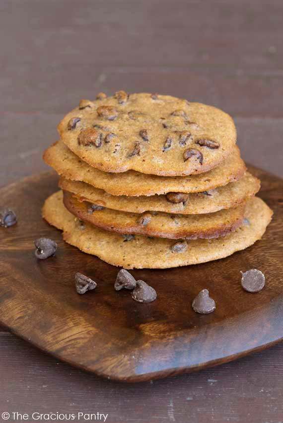 5 Clean Eating Spelt Chocolate Chip Cookies stacked on a small, square, dark wood cutting board with rounded edges. There are a few chocolate chips scattered around the base of the cookies.