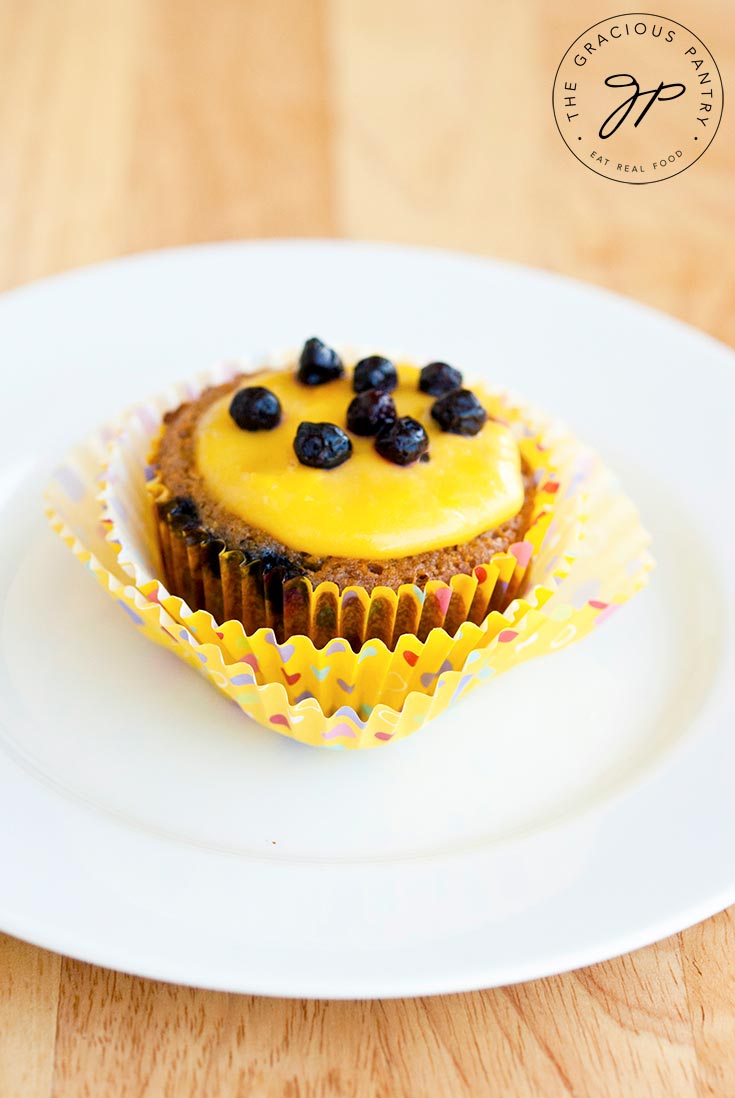 A single cupcake from a batch of these Clean Eating Blueberry Cupcakes with Lemon Curd Frosting sits on a small, white plate. The cupcake paper is pulled away from the cupcake and the lemon curd frosting on top of the cupcake is topped with blueberries.