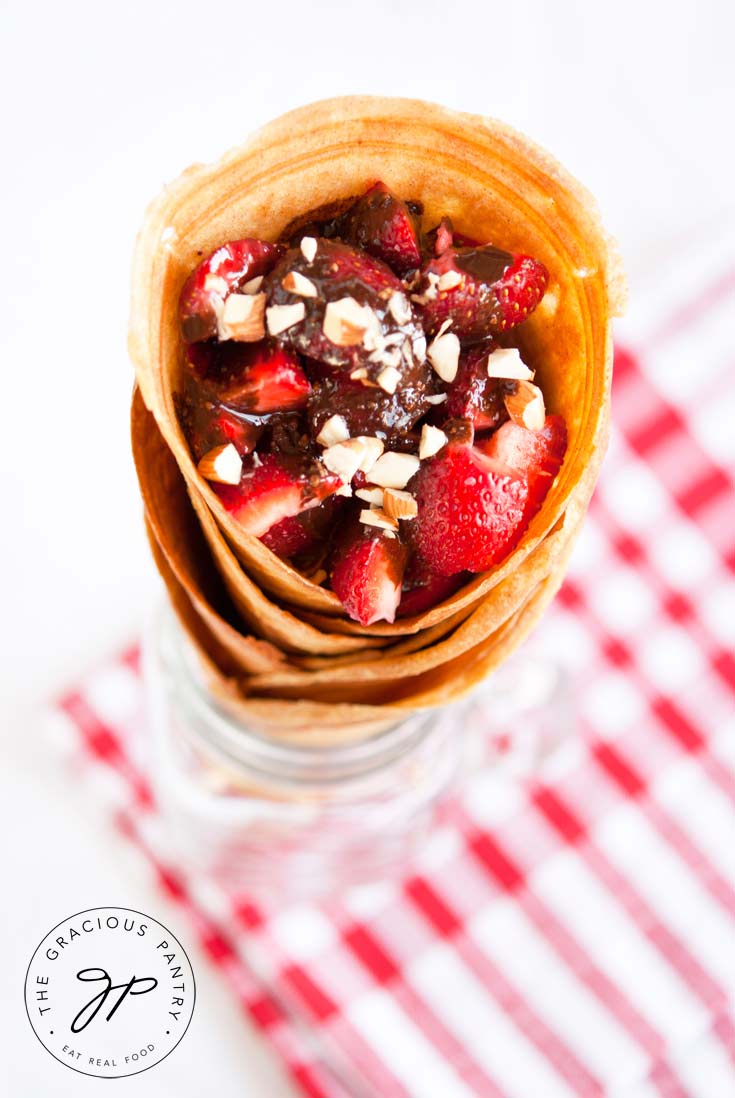A view of a stack of Clean Eating Waffle Cones, from almost the top looking down at an angle. The top cone is filled with cut strawberries that have been drizzled with chocolate and sprinkled with almond pieces. The stack of cones sits in a glass mason jar mug.