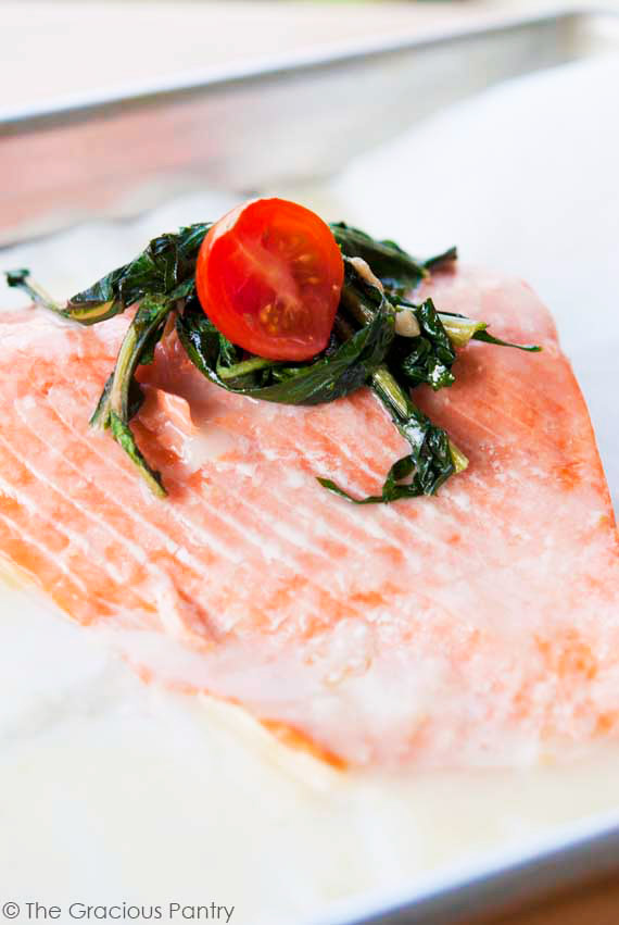 This Clean Eating Baked Salmon With Roasted Garlic Lemon Sauce sits on the silver baking pan it was cooked on. The fillet is topped with a small bit of greens and half a cherry tomato for garnish.