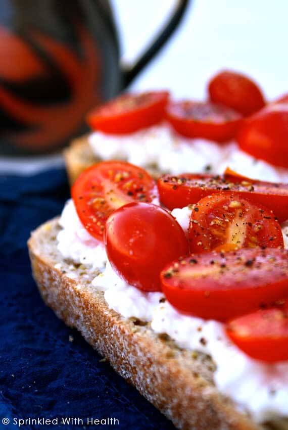 Two slices of toast sit on a dark surface, they are topped with cottage cheese and halved cherry tomatoes.