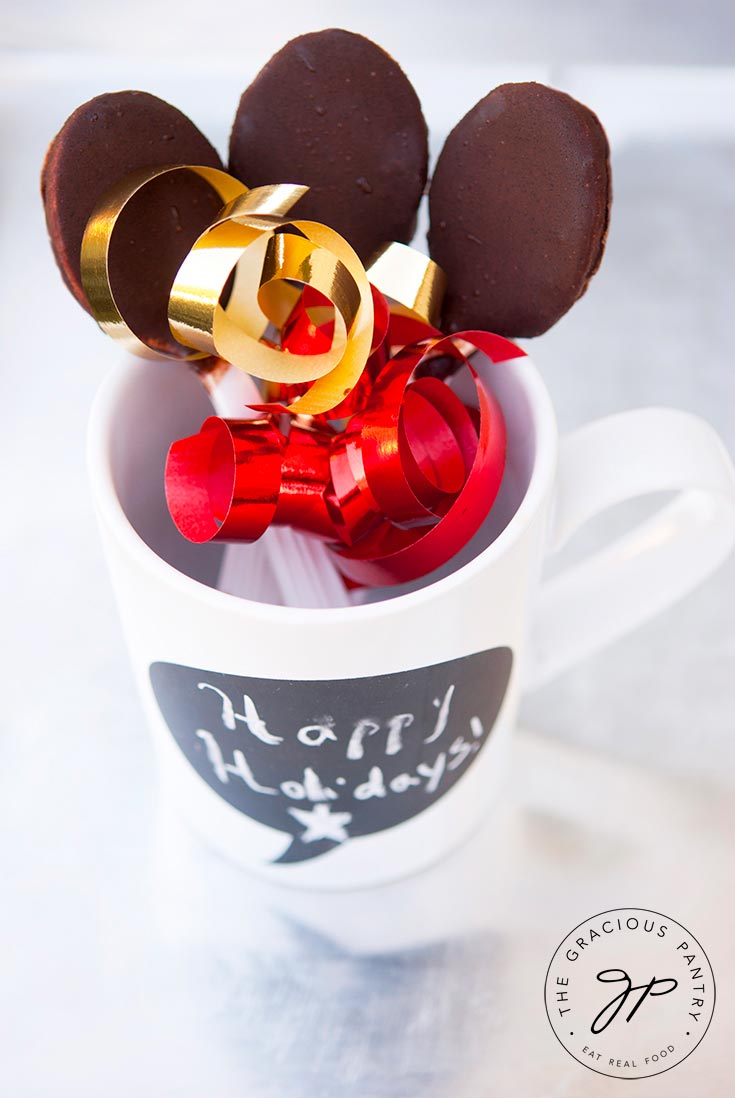 Three Clean Eating Chocolate Covered Spoons are held in a white mug with the chocolate end up. Colorful gold and red ribbon has been tied around them for a festive look.