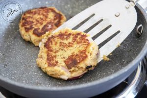 Two clean eating tuna patties cooking in a skillet as one gets flipped over with a metal spatula.