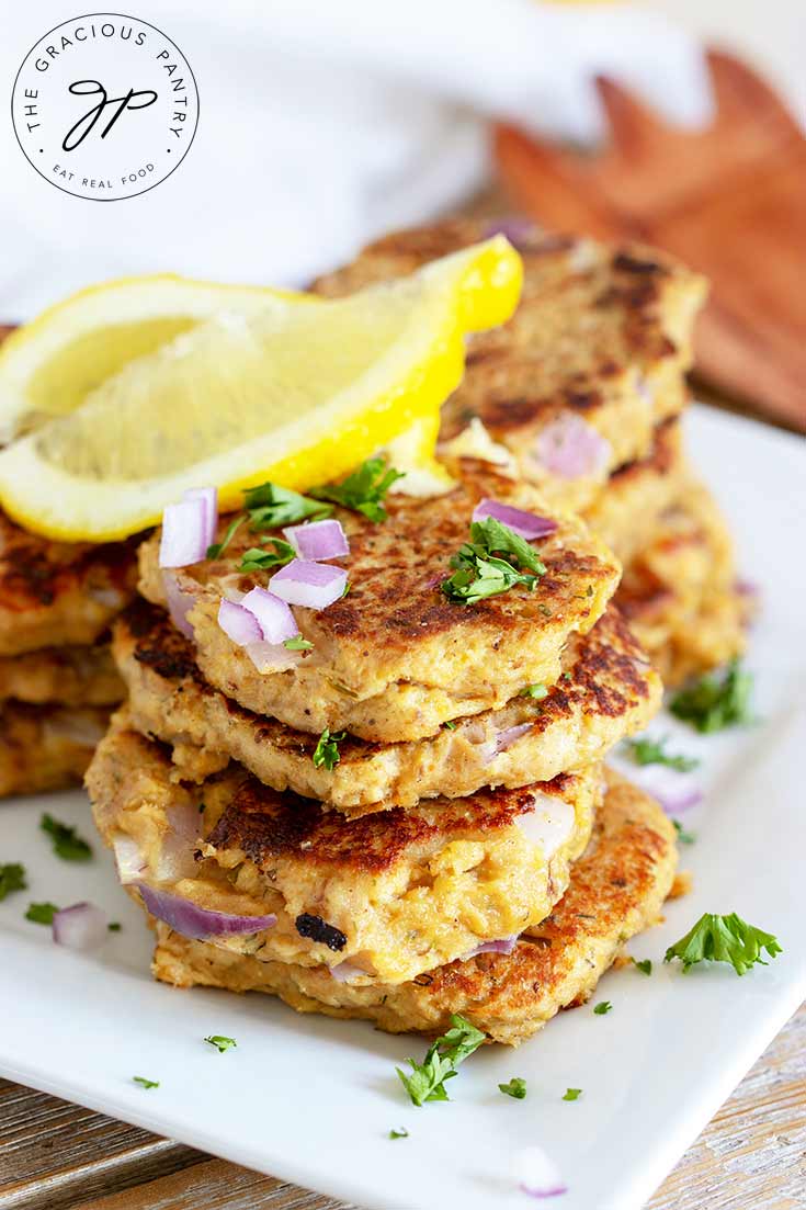 A stack of theseClean Eating Tuna Patties sits on a white plate topped with fresh herbs, chopped red onion and some bright yellow lemon wedges.