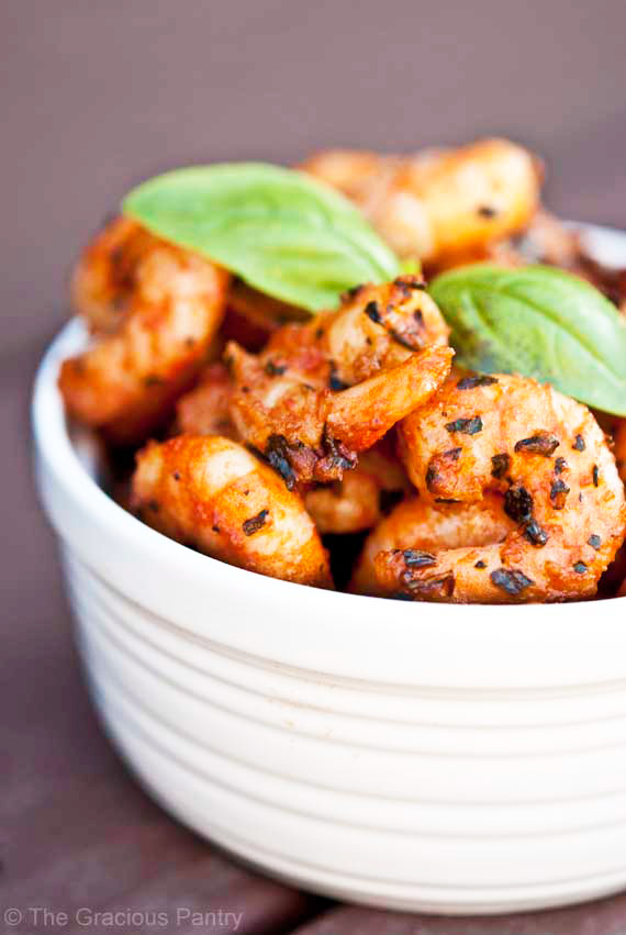 Italian Shrimp piled high in a white bowl. The large shrimp are shown up close and you can see the herbs on them. There is a fresh basil leaf on top of the shrimp for garnish.