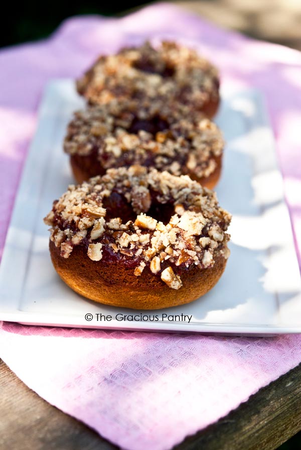 There are three Clean Eating Doughnuts signed up on a long, rectangular, white platter. The platter sits on a pink cloth napkin which sits on a wooden table. The doughnuts are lined up from front to back and you can see the red fruit spread with sprinkled nuts over the top.