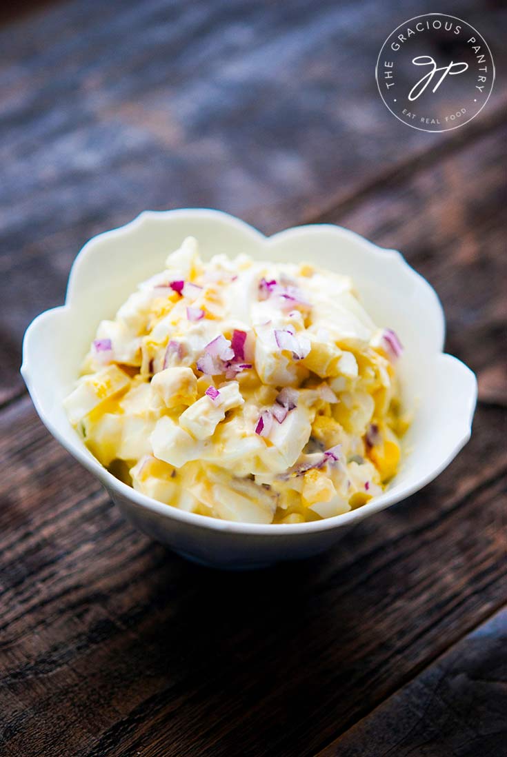 A white, fluted bowl sits filled with this clean eating egg salad recipe on a wooden background. You can see bits of chopped red onion mixed into the bright yellow and white egg salad.