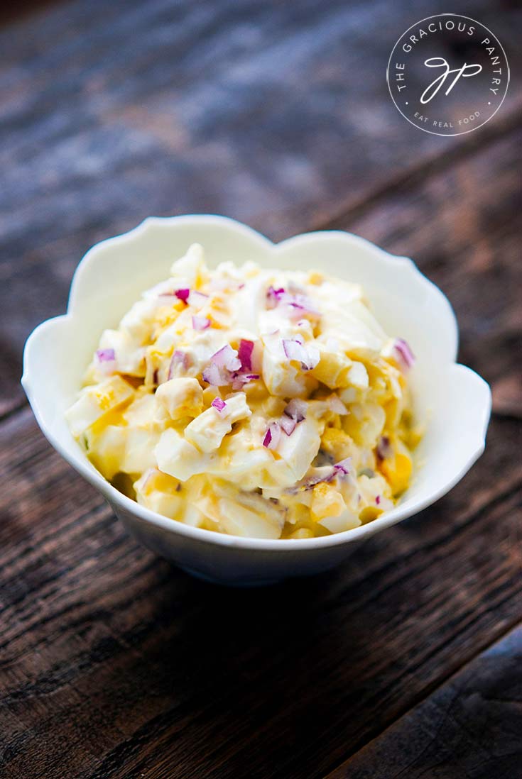A white, fluted bowl sits filled with this clean eating egg salad recipe on a wooden background. You can see bits of chopped red onion mixed into the bright yellow and white egg salad.