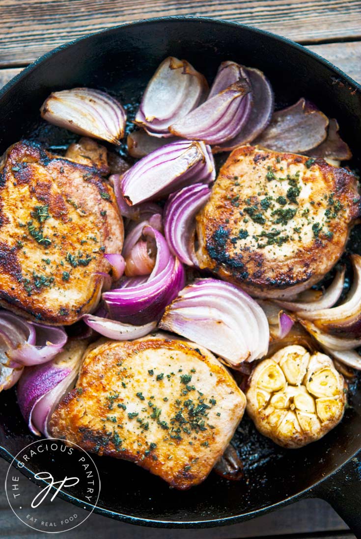 Clean Eating Roasted Garlic And Onion Pork Chops shown in the skillet they were cooked in. There are three perfectly browned pork chops with wedges of roasted red onion and a roasted head of garlic.