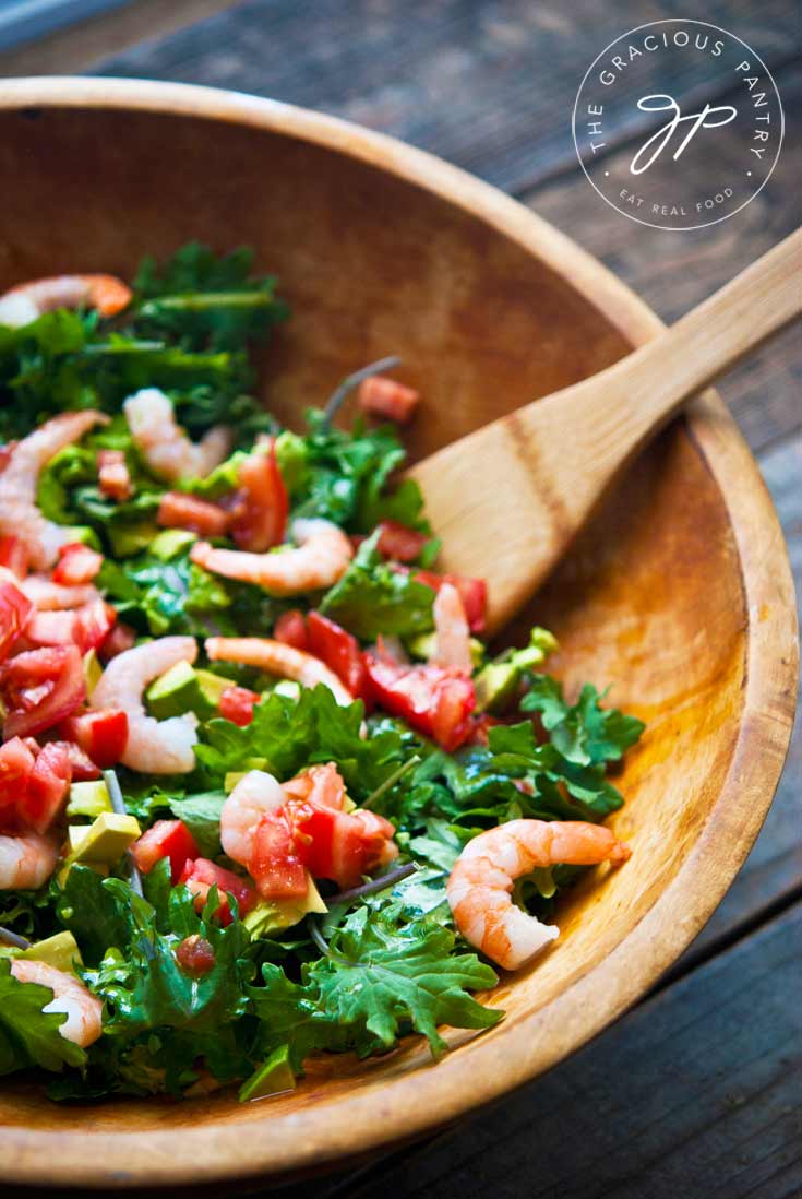 A close up shot of this Clean Eating Shrimp And Kale Salad in a large wooden bowl. You can see the greens at the bottom are topped with bright colored shrimp and red tomatoes.