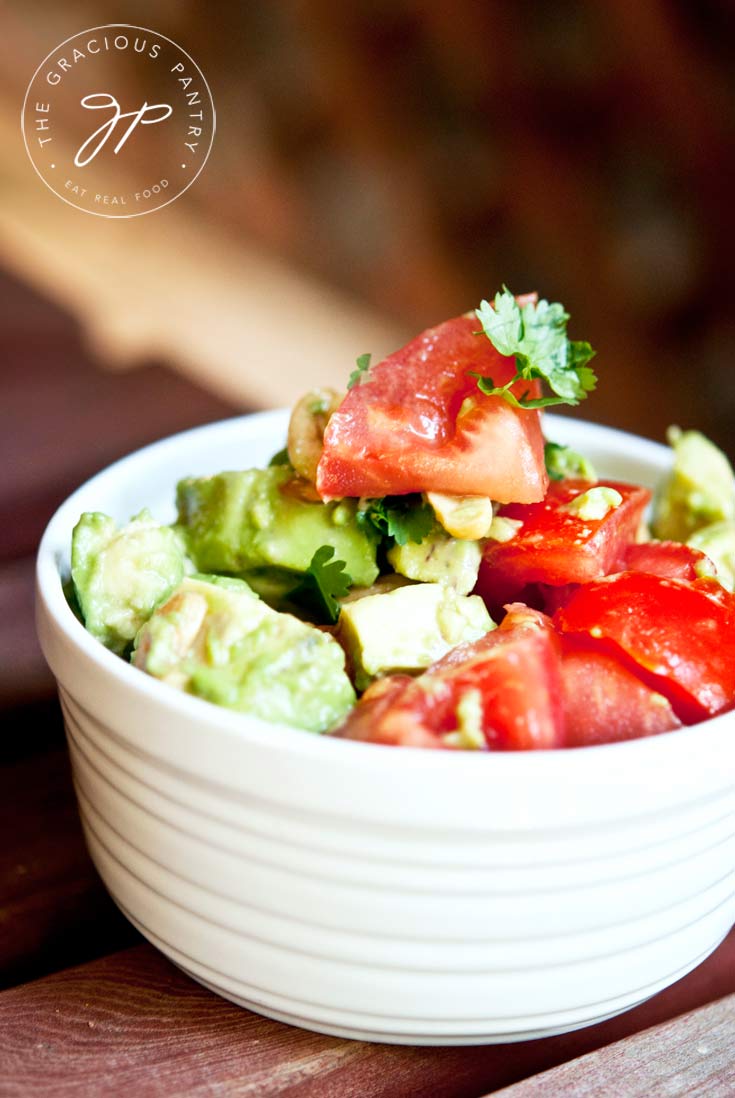 A close up view of a white bowl filled with this Clean Eating Avocado Cashew Salad. You can see chunks of avocado and tomatoes over the top of the bowl.