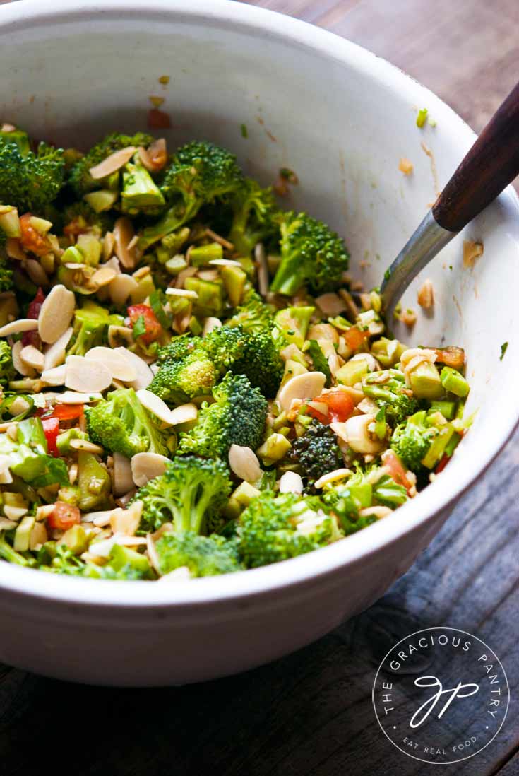 A large white bowl sits filled half way with this Clean Eating Broccoli Salad. Salad spoons are in the bowl as well for serving. You an see tons of broccoli, lots of sliced almonds and a few hints of chopped tomato.