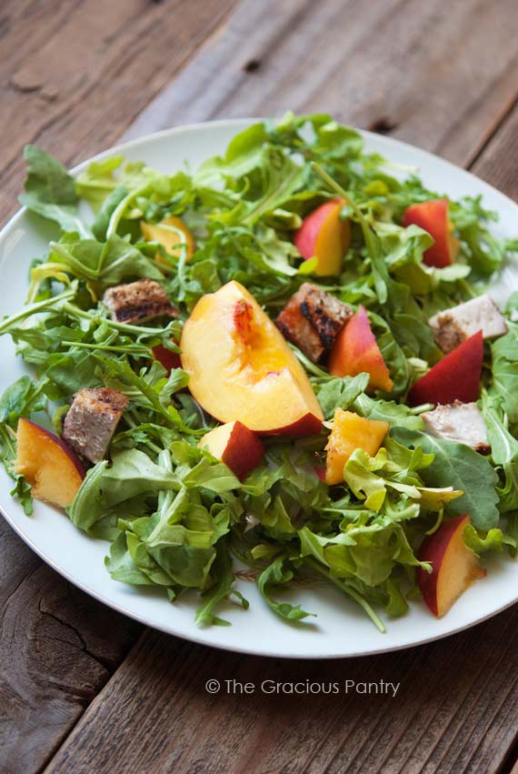 Clean Eating Pork Peach Arugula Salad With Peach Vinaigrette With Peach Vinaigrette sits on a white, round plate, ready to eat. The peaches and pieces of pork sit on a large bed of green arugula.