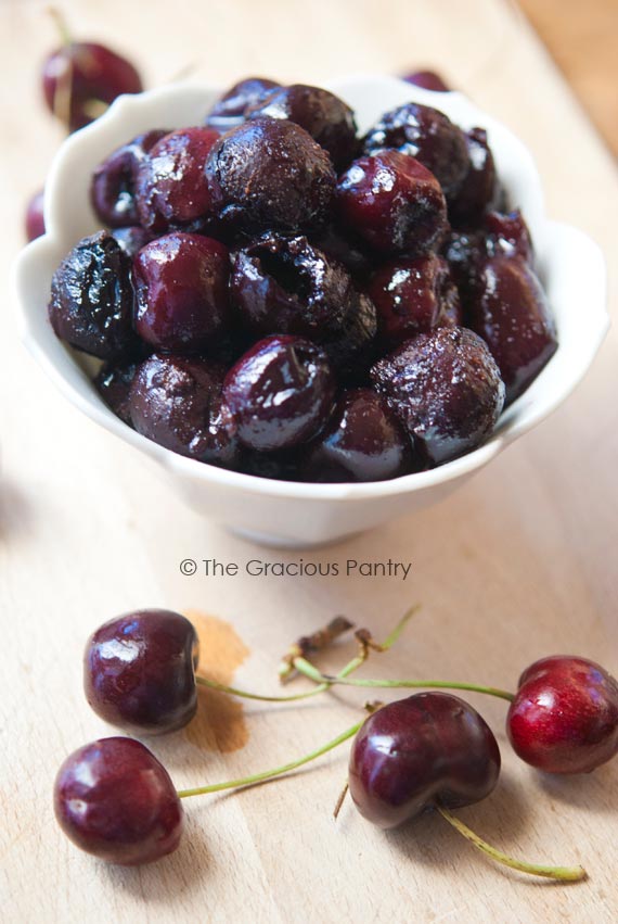 Clean Eating Barbecued Fresh Cherries sitting in a white bowl with fresh, uncooked cherries laying around the base of the bowl.