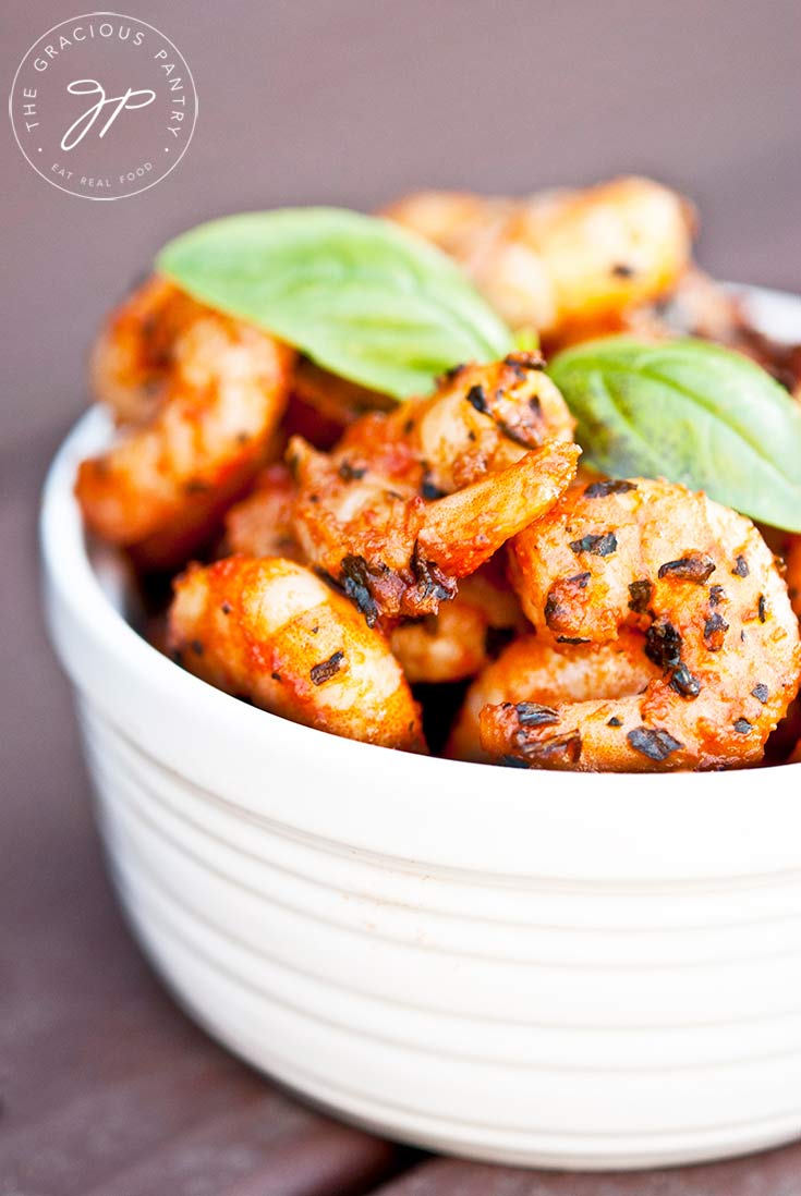 A white bowl filled with these delicious Clean Eating Italian Style Shrimp and topped with a fresh basil leaf. The shrimp are a bright orangey-red color and you can see the blackened spices clinging to the shrimp.