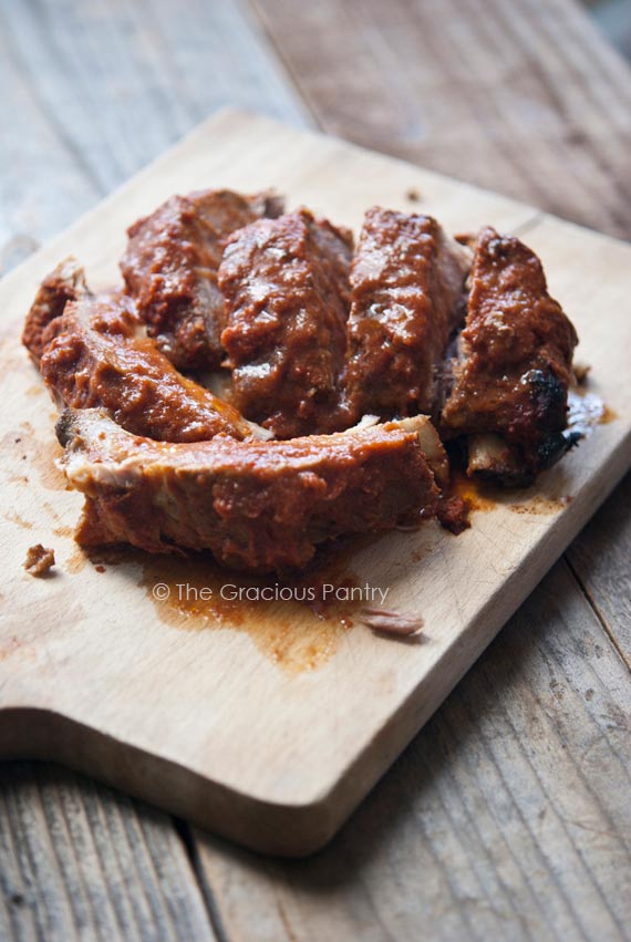 A small pile of freshly made, Clean Eating Slow Cooker Baby Back Ribs piled on a cutting board with rib sauce all over them.