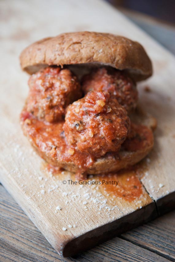 A single Clean Eating Spaghetti Meatball Sandwich sits on a cutting board. There are three large meatballs between a whole grain bun. The top half of the bun has been set back a bit so you can see more of the meatballs. The meatballs are covered in marinara.