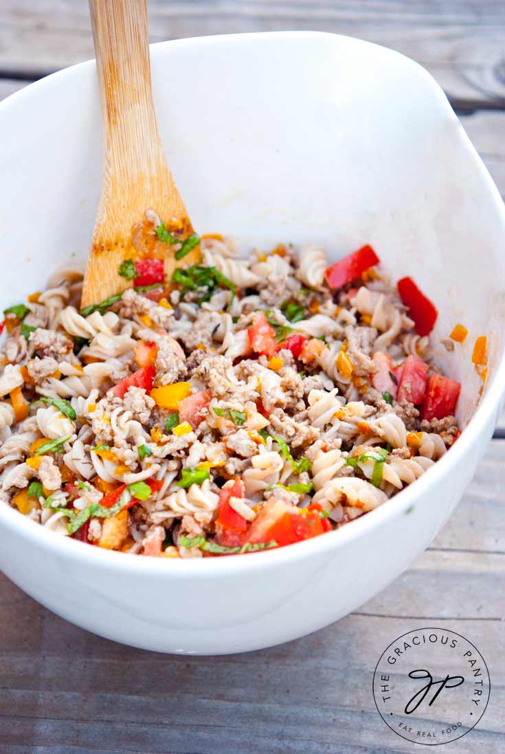 A large, white serving bowl sits filled with this Clean Eating Turkey Vegetable Pasta Salad. A wooden spoon rests on the side of the bowl. The pasta is colorful with all the flecks of brightly colored vegetable tossed about in the bowl.