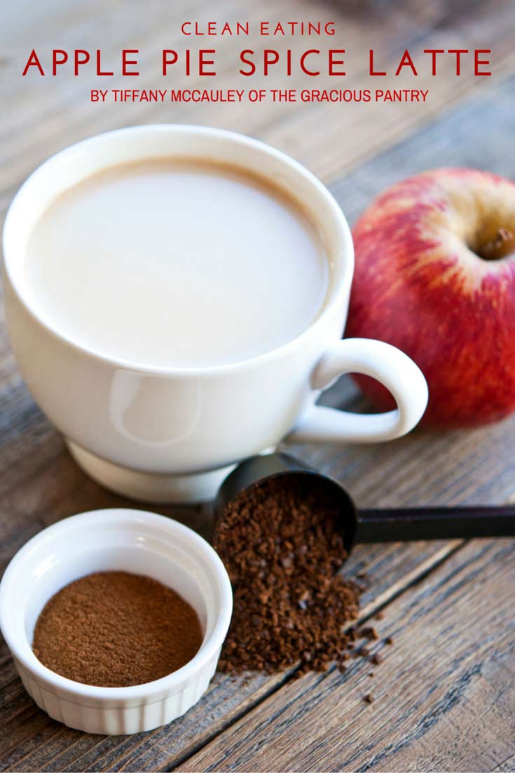 a white mug filled with Clean Eating Apple Pie Spice Latte sits on a wooden table. Next to the mug is an apple, a coffee scoop and a small white dish with coffee grounds.