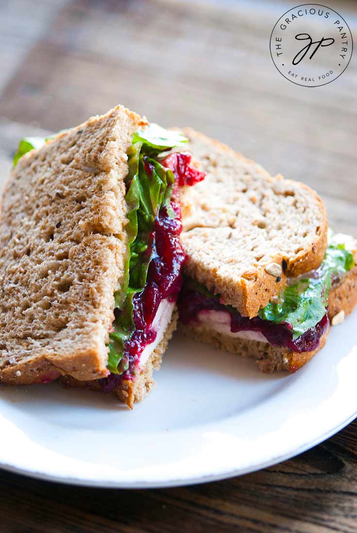 A delicious Leftover Turkey Sandwich sits on a plate, ready to sink your teeth into. There are layers of bread, turkey, cranberry sauce and lettuce visible.