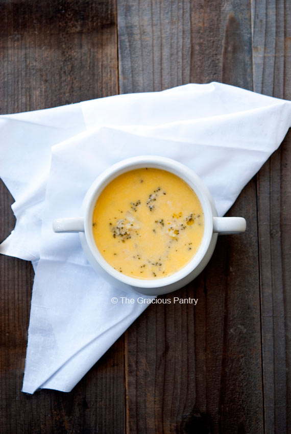 An overhead view looking down into a white bowl filled with bright, golden yellow, Clean Eating Broccoli Cheddar Soup. A white napkin sits unfolded to the side.