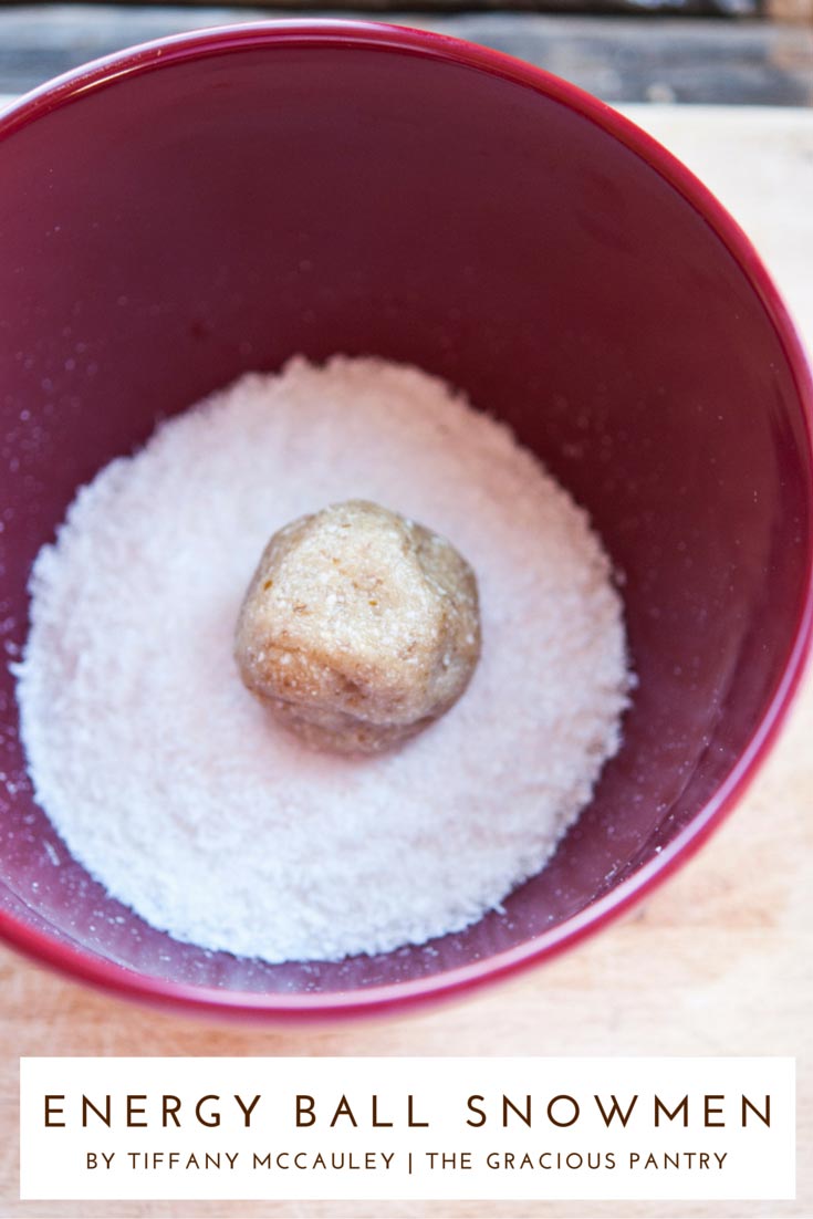 A single energy ball sits in a bowl of coconut flakes, waiting to be rolled in it to construct one of these Clean Eating Energy Ball Snowmen.