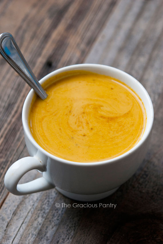 A white mug on a wooden table, filled with Curried pumpkin soup.