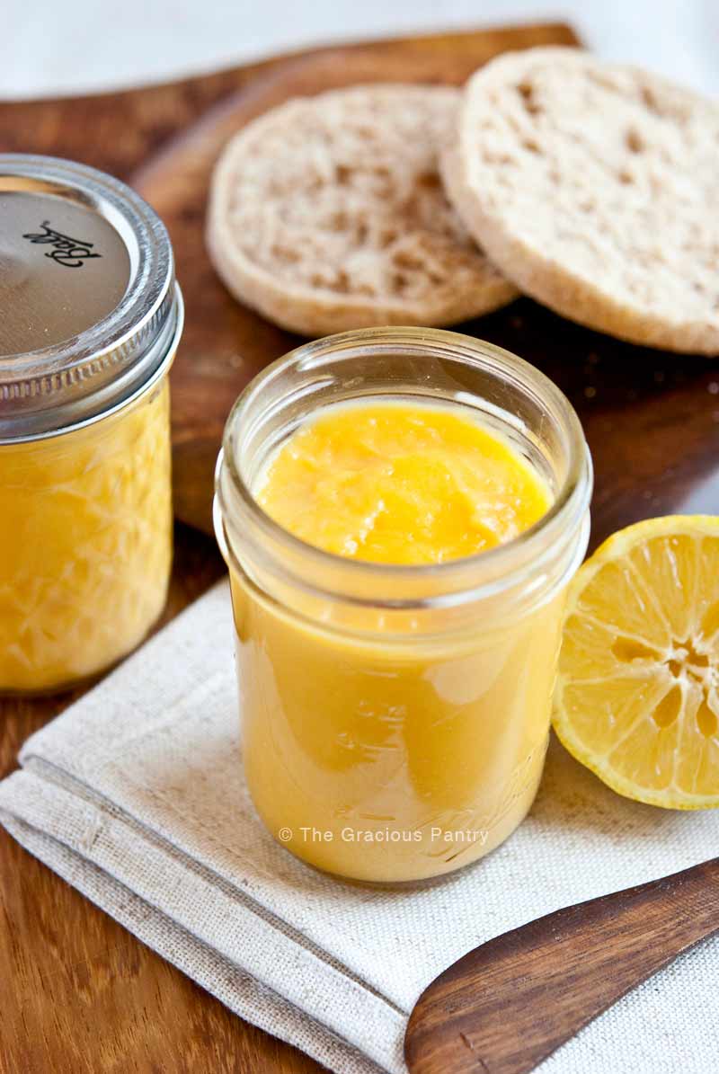 Two pint-sized canning jars of homemade lemon curd recipe, surrounded by lemons and whole grain English muffin