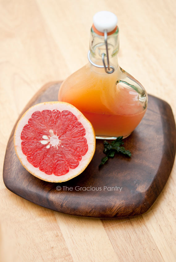 Clean Eating Grapefruit Vinaigrette Recipe shown in a dressing bottle sitting next to half a grapefruit. Both are sitting on a small, square, dark wood cutting board with rounded corners.