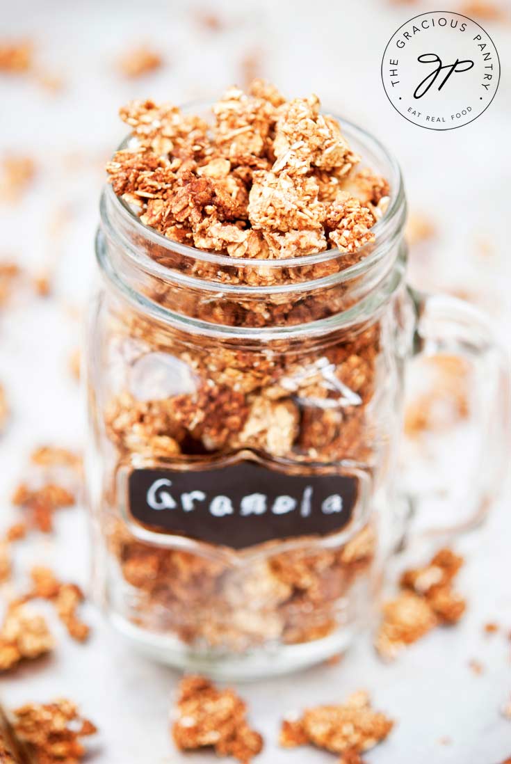 A mason jar sits filled to the rim with Clean Eating Maple Cinnamon Granola. There are small chunks of granola scattered around the base of the jar as well.