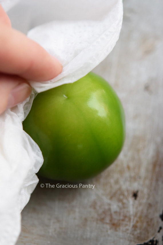 Cleaning your tomatillos is critical. Image shows the sticky coating of the tomatillo being wiped off with a white cloth and some vinegar.