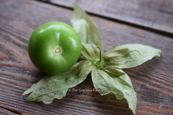 Image of a tomatillo with the husk removed and sitting to the side of it. The bright green tomatillo sits just to the left of the husk which has been flattened out like a star.
