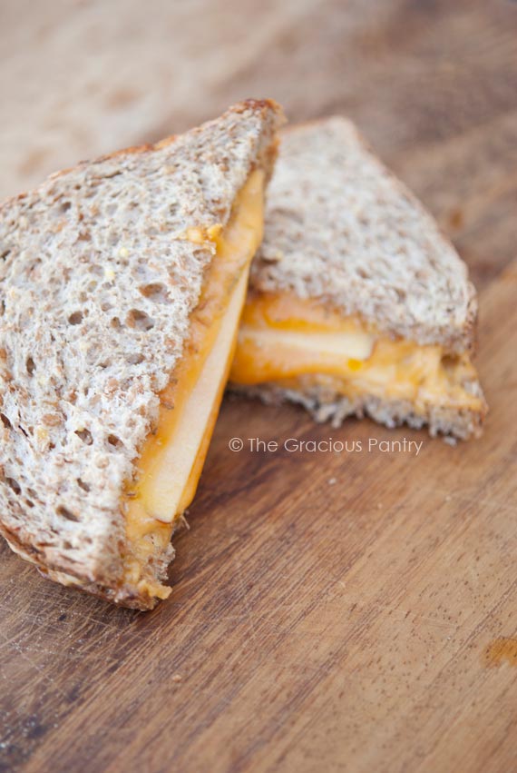 A Campfire Grilled Cheese Sandwich with apples and thyme, lays cut in half on a wood cutting board.