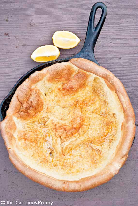 Clean Eating Dutch Baby Recipe shown from overhead looking down into the pan which is still in the skillet it was baked in. Two lemon wedges sit on the table just outside the black skillet. The dutch baby is golden brown and sprinkled with unprocessed sugar and lemon juice.