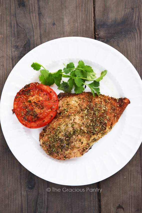 Clean Eating Roasted Balsamic Chicken And Tomatoes shown from overhead on a white plate. One chicken breast and one half of a roasted tomato sit on the plate with a small amount of fresh greenery on the plate for garnish.
