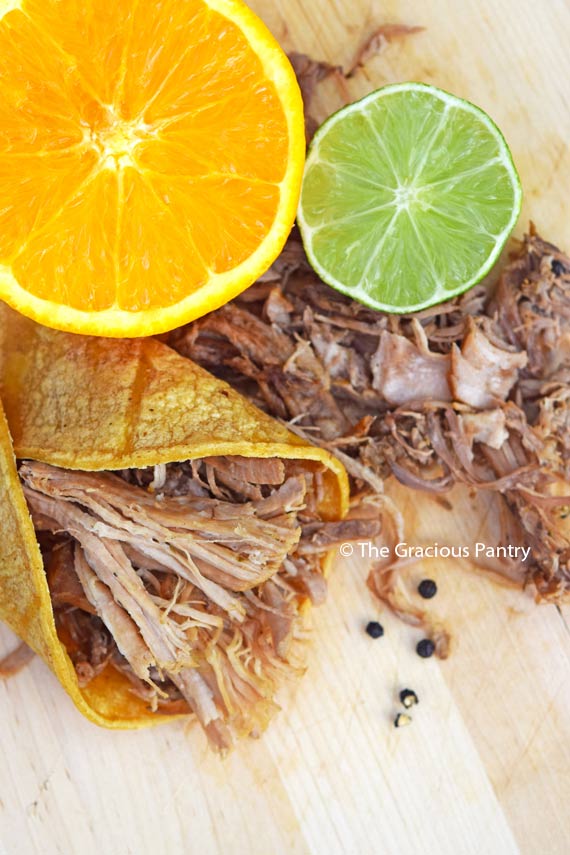 Clean Eating Slow Cooker Carnitas Recipe, shredded and displayed wrapped in a tortilla next to a halved orange and lime. Black peppercorns are roughly ground and sprinkled over the carnitas.