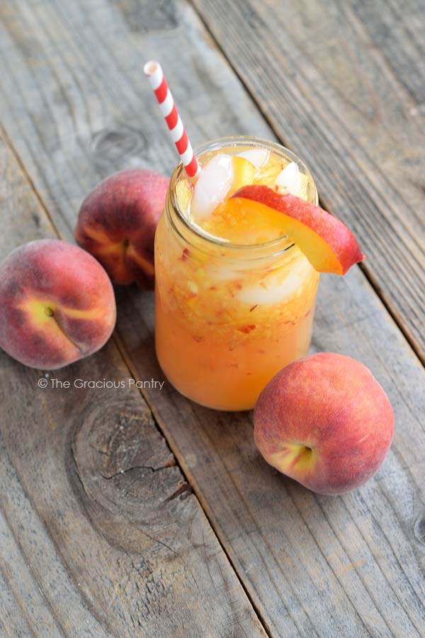 A glass of Clean Eating Georgia Peach Lemonade sits on a wooden table. It has a red and white stripped straw in the clear, glass mason jar mug and there are three peaches around the base of the mug on the table.