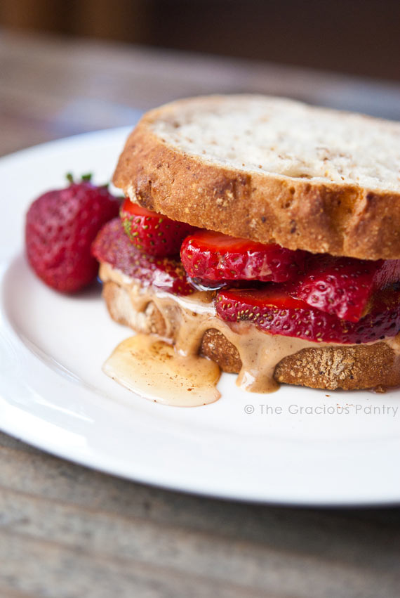 A Clean Eating Fruit And Nut Butter Sandwich sits on a white plate. You can see the sliced strawberries between the slices of bread and the honey and peanut butter oozing over the edge onto the plate just a tiny bit.