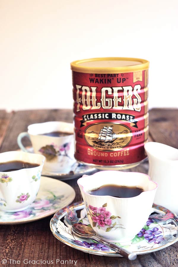 Three teacups on saucers sit around a can of Folger's coffee filled with this Oma's Holiday Spiced Coffee recipe.