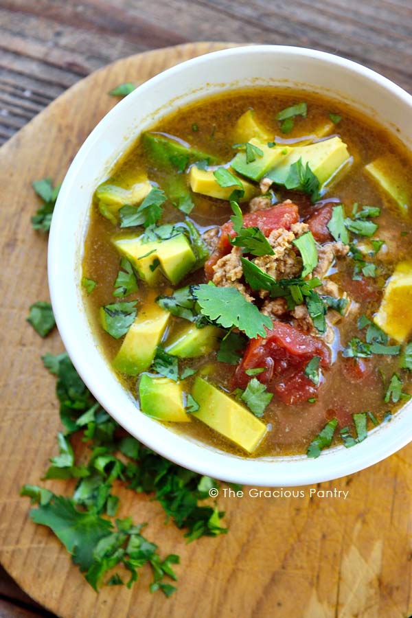 Clean Eating Slow Cooker Taco Soup in a white bowl sitting slightly off to the right. Shows the soup from the top including meat, chopped avocado and tomatoes with fresh cilantro sprinkled over the top. 