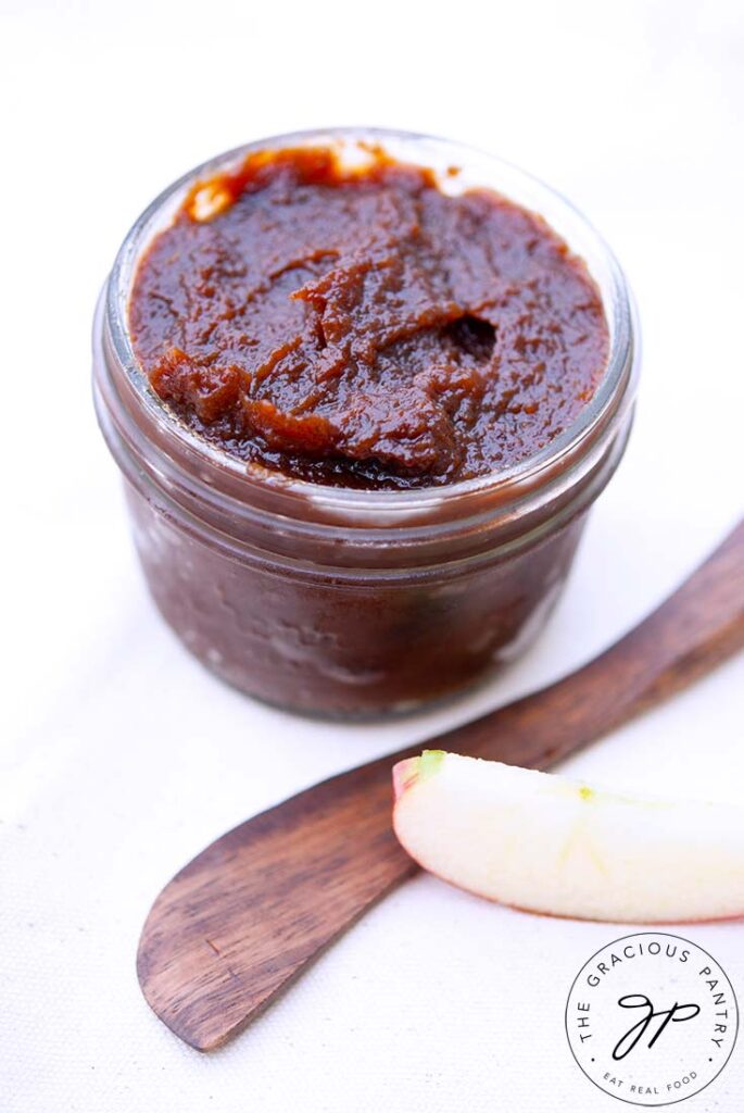 An open jar of this crock pot apple butter sits next the a butter knife and a slice of apple.