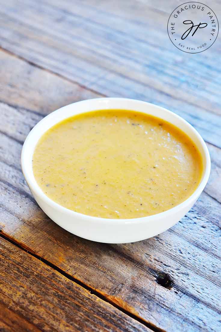 A white, round bowl sits on a wooden table filled with this reddish-gold colored, clean eating roasted red pepper bisque.