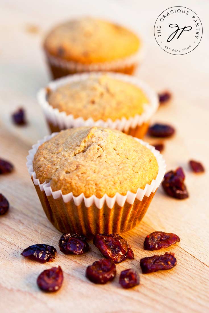 Three Clean Eating Cranberry Oat Bran Muffins are lined up on a wooden surface. They have golden brown tops and are in white cupcake liners.