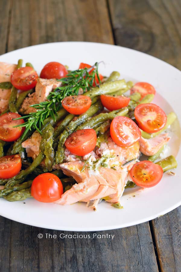 Clean Eating Instant Pot Mediterranean Rosemary Salmon broken into pieces and topped with asparagus, cherry tomatoes, a sprig of fresh rosemary and finally drizzled with a bit of fresh olive oil.