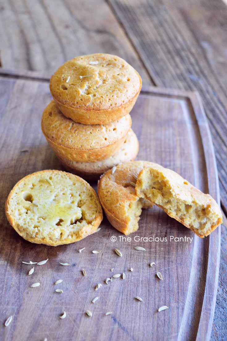 These Clean Eating Almond Fennel Muffins are perfect for the spring holidays! There are three stacked up here on a cutting board with half a sliced muffin in front of the stack and a broken muffin just to the right of that.