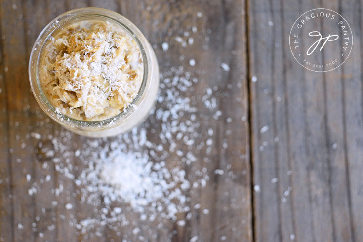 Clean Eating Coconut Bomb Oatmeal Recipe shown from overhead looking down into the jar of oats. Coconut flakes are sprinkled on the oats as well as on the wood table the jar is sitting on.