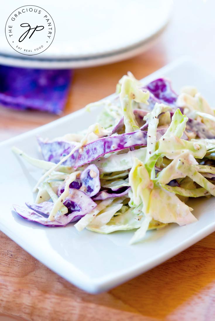 Clean Eating Coleslaw on a white serving plate, with a stack of two white plates sitting behind it. The coleslaw shows strips of both green and red cabbage.