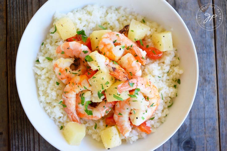 Clean Eating Sheet Pan Pineapple Shrimp Recipe shown from overhead. Presented in a white bowl and showing the entire dish of shrimp, pineapple chunks and green garnish over a bed of riced cauliflower.